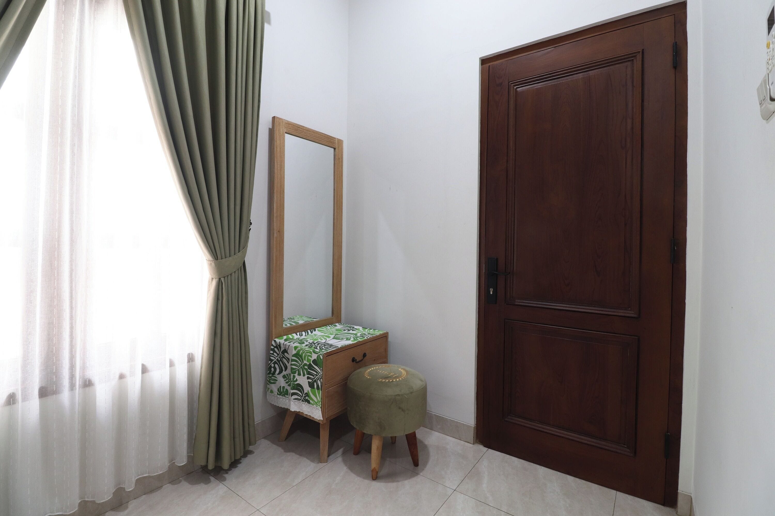 A room with a wooden door, a mirror, a small stool, and a window with curtains.