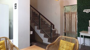 A staircase with a glass railing, a wooden table, wicker chairs with yellow cushions, and a wall with a green textured backdrop and a sign. - Bisma Balcony Homestay (Pogung Lor)
