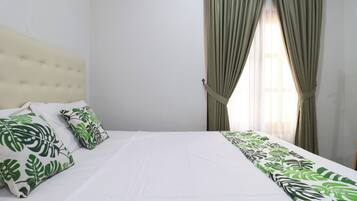 Una cama bien hecha con colcha blanca y almohadas estampadas con hojas, un cabecero capitonado y una ventana con cortinas.