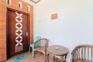 Una habitación con puerta de madera con diseño geométrico, una pequeña mesa redonda, dos sillas de madera y una silla de plástico azul, además de un felpudo verde.