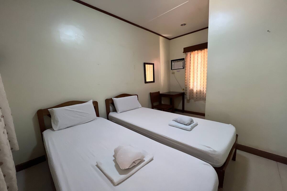 Habitación de hotel con dos camas individuales, cabecero de madera, un escritorio pequeño y una ventana con cortinas.