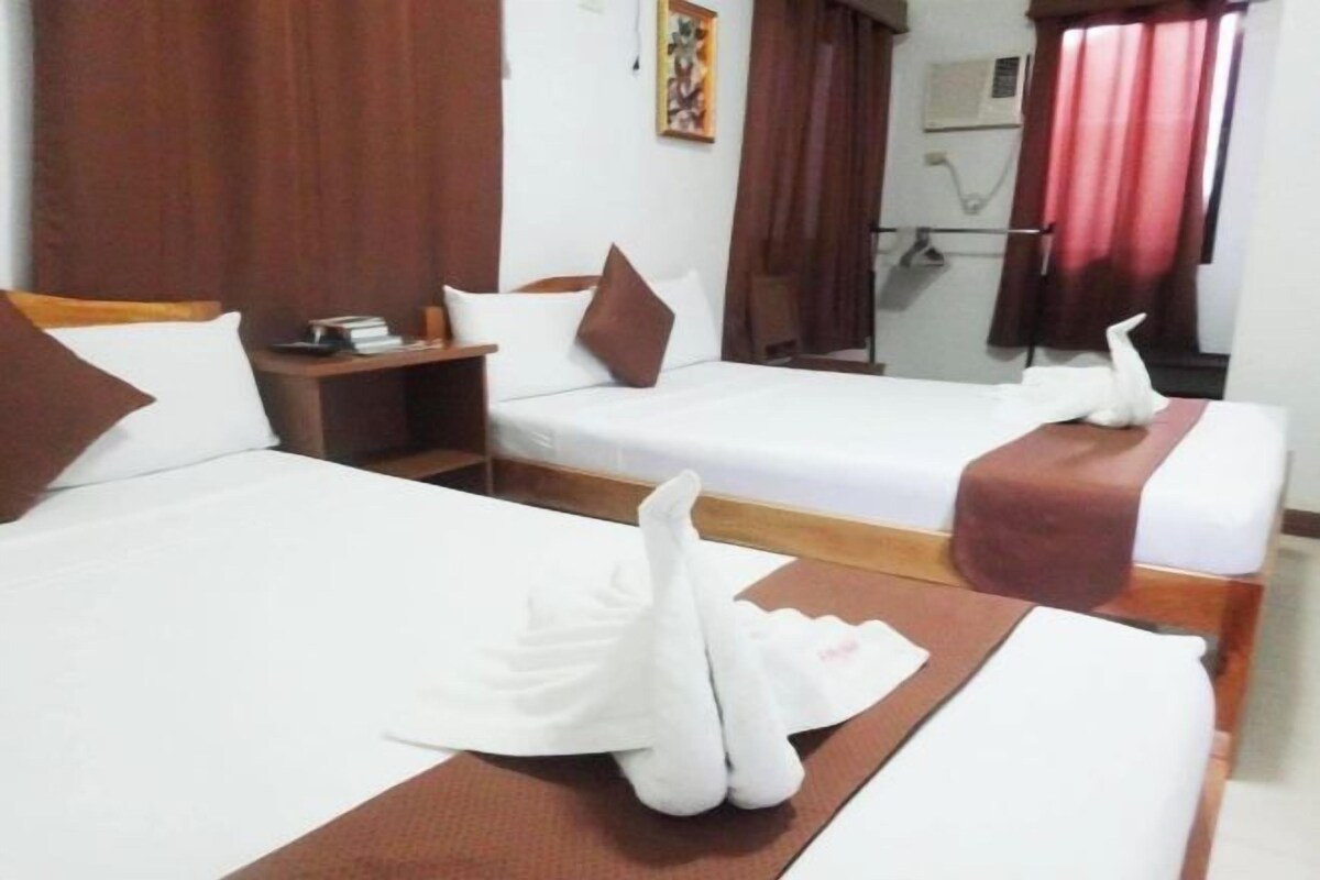 Habitación de hotel con dos camas, cada una con ropa blanca y almohadas marrones. Hay una mesita de noche de madera entre las camas, y una pequeña mesa con un libro y un teléfono. La habitación tiene un cabecero de madera, una ventana con cortinas y un aire acondicionado instalado en la pared.