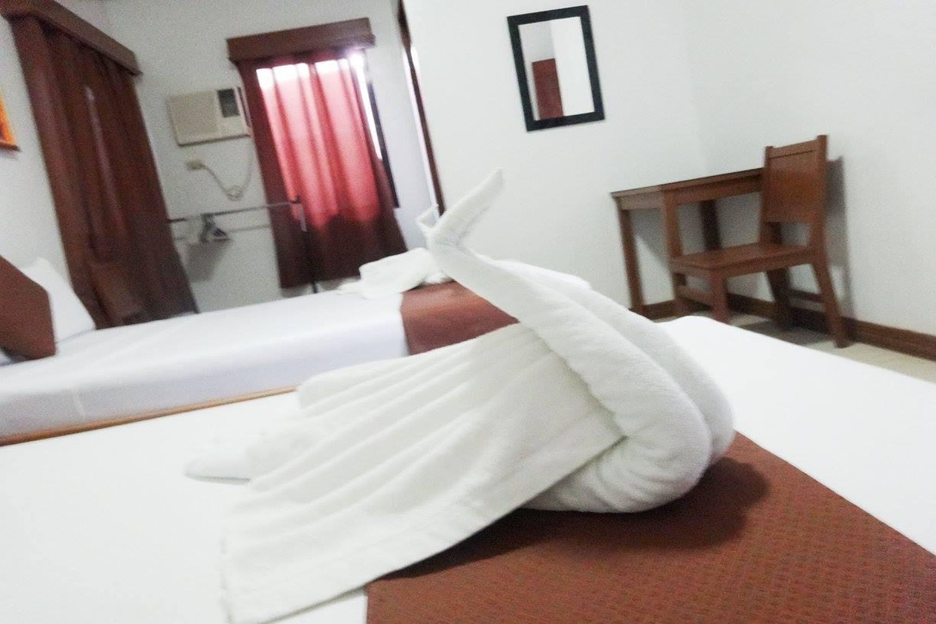 Una habitación de hotel con una cama, una silla de madera, un espejo y una ventana con cortinas rojas.