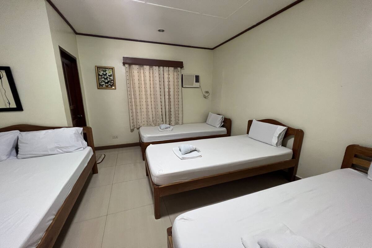Una habitación de hotel con tres camas individuales, una ventana con cortinas, un aire acondicionado montado en la pared y un cuadro enmarcado.