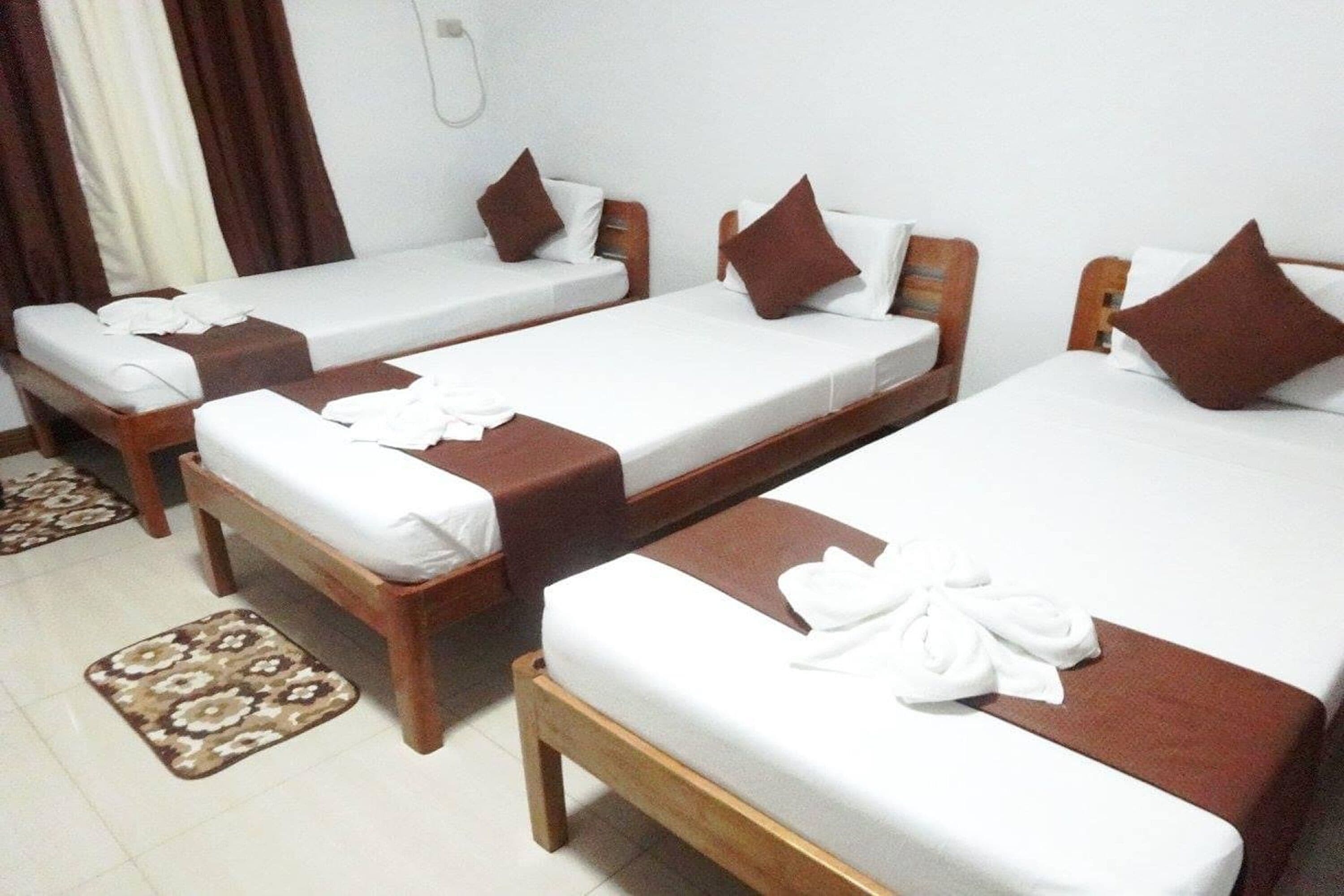 A hotel room with three single beds, each with a wooden frame and white bedding. There are brown pillows on each bed, and a brown bed runner. The room has a tiled floor and a patterned rug in front of the beds.