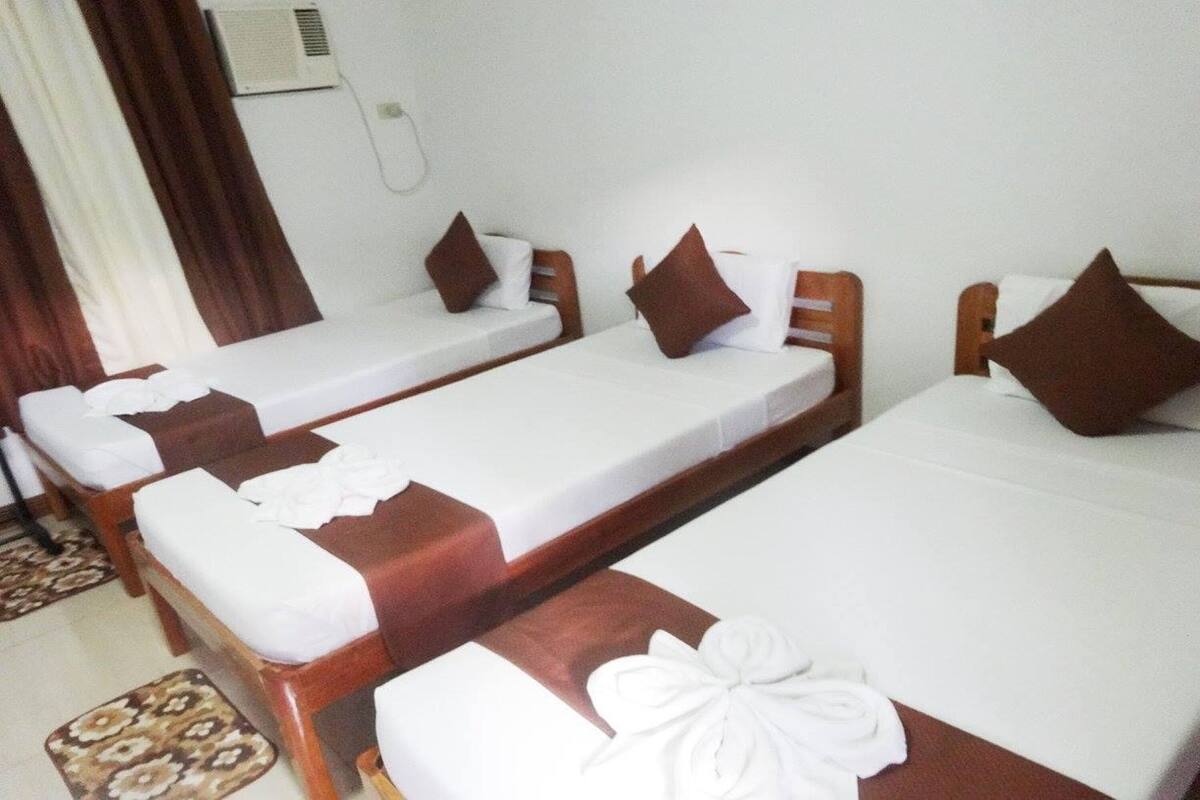 Habitación de hotel con tres camas individuales, cada una con cabecero de madera y ropa de cama blanca. Hay una alfombra pequeña en el suelo y un aire acondicionado instalado en la pared.