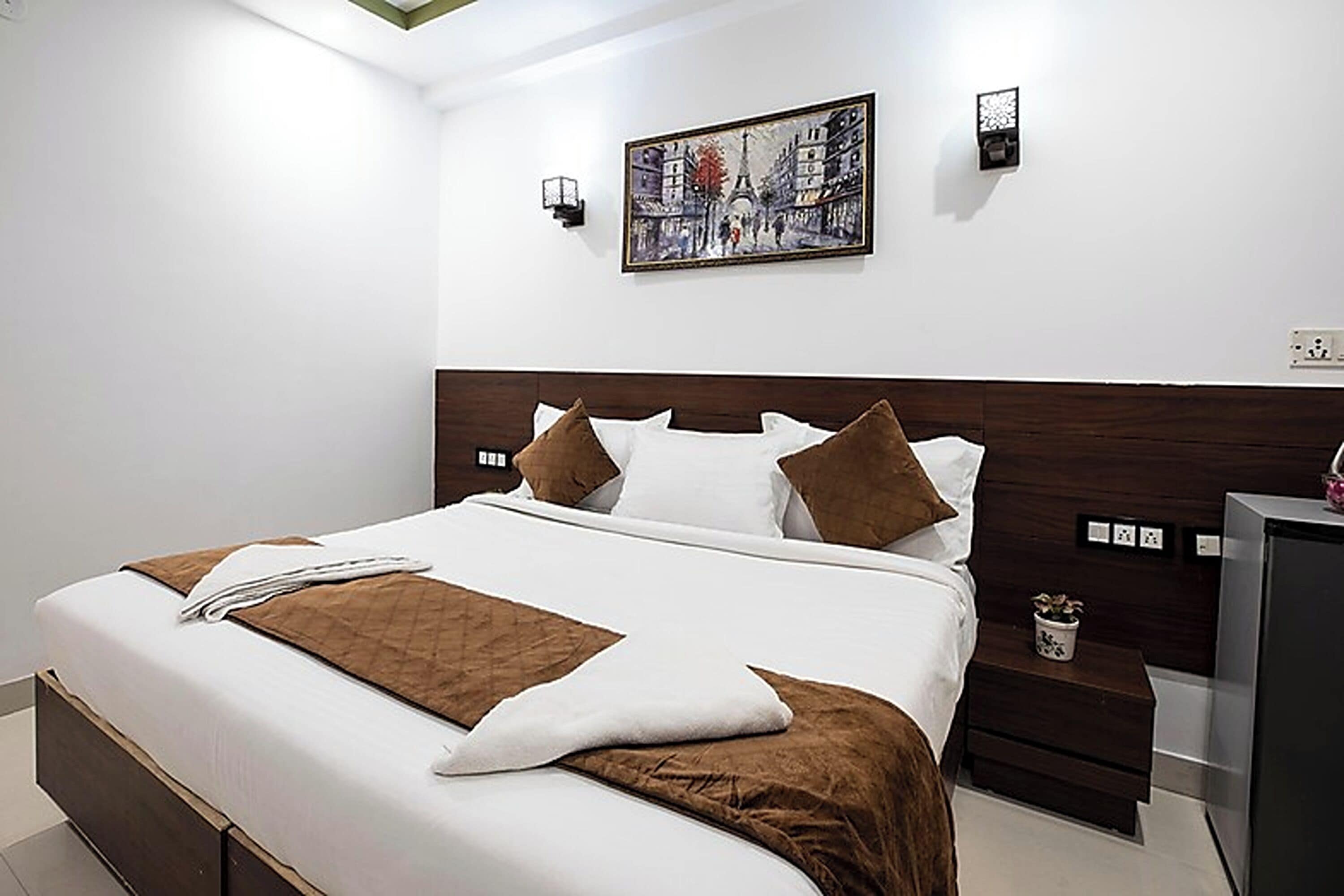 Una cama bien hecha con colcha marrón y almohadas, una mesita de noche con una planta y un cuadro de un paisaje urbano en la pared.
