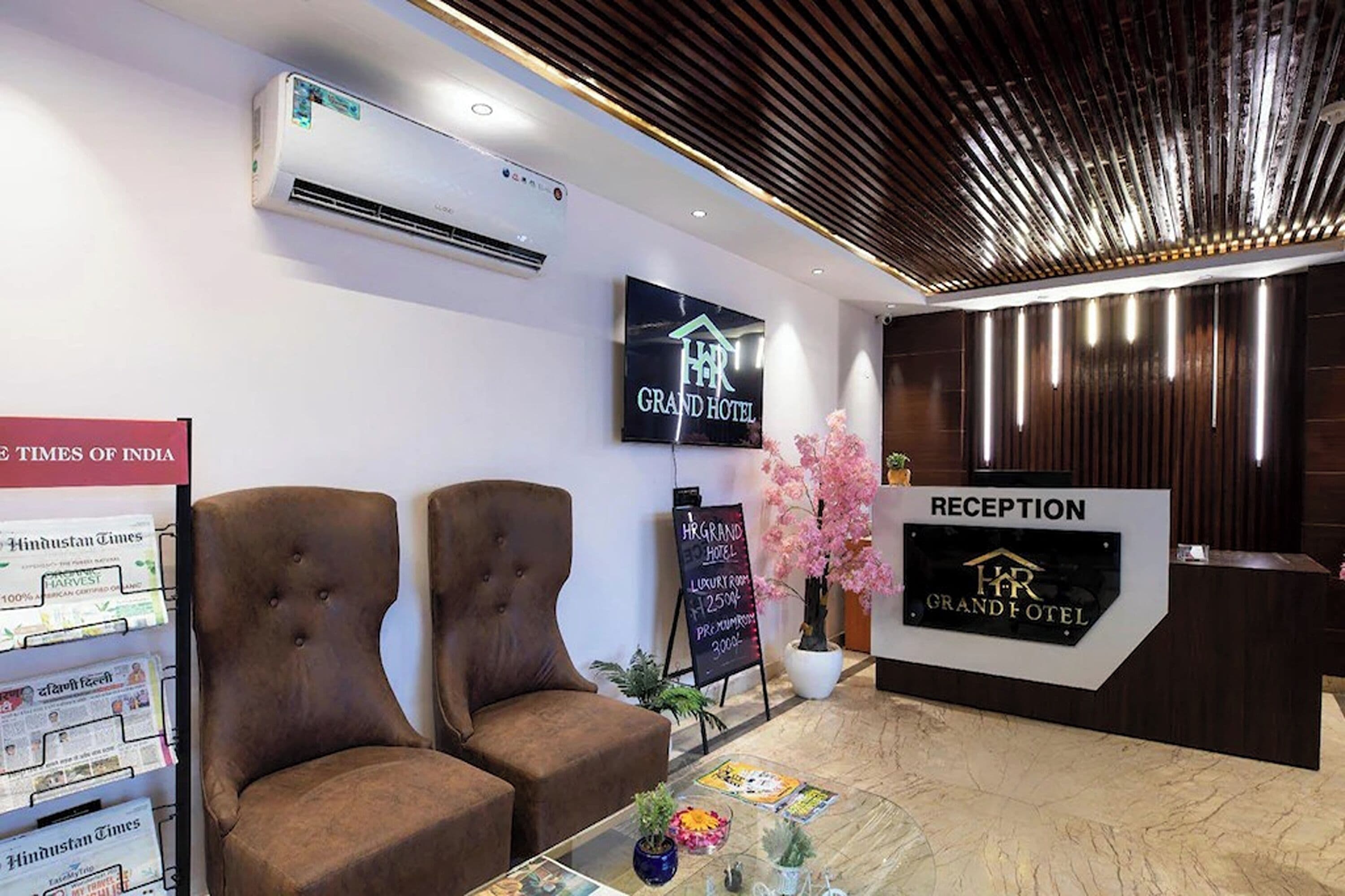 A hotel reception area with a reception desk, two brown upholstered chairs, a wall-mounted air conditioner, and a sign for the HR Grand Hotel.