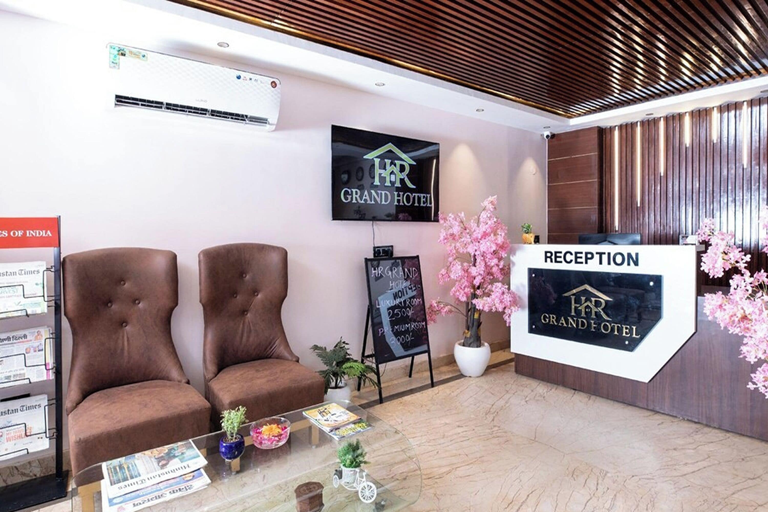 A hotel reception area with a reception desk, two brown upholstered chairs, a potted plant, a glass table with magazines, and a sign indicating the reception.
