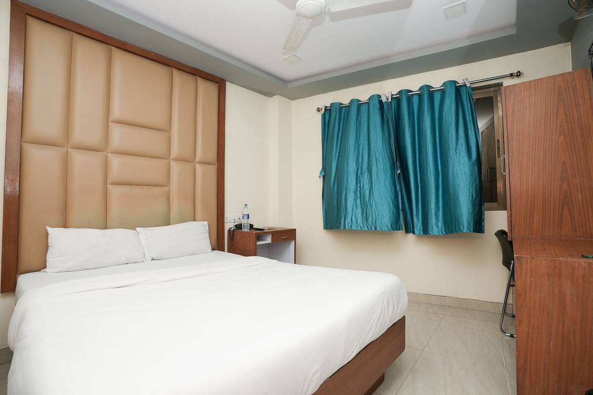 A hotel room with a bed, a wooden headboard, a desk with a chair, a wardrobe, and teal curtains.