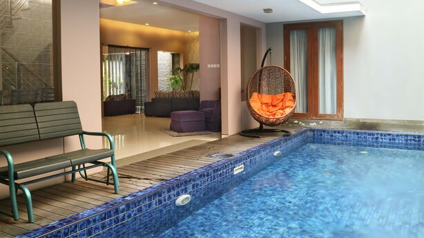 Una piscina interior con un banco y una silla colgante, una escalera y una sala de estar con sofá y cortinas.