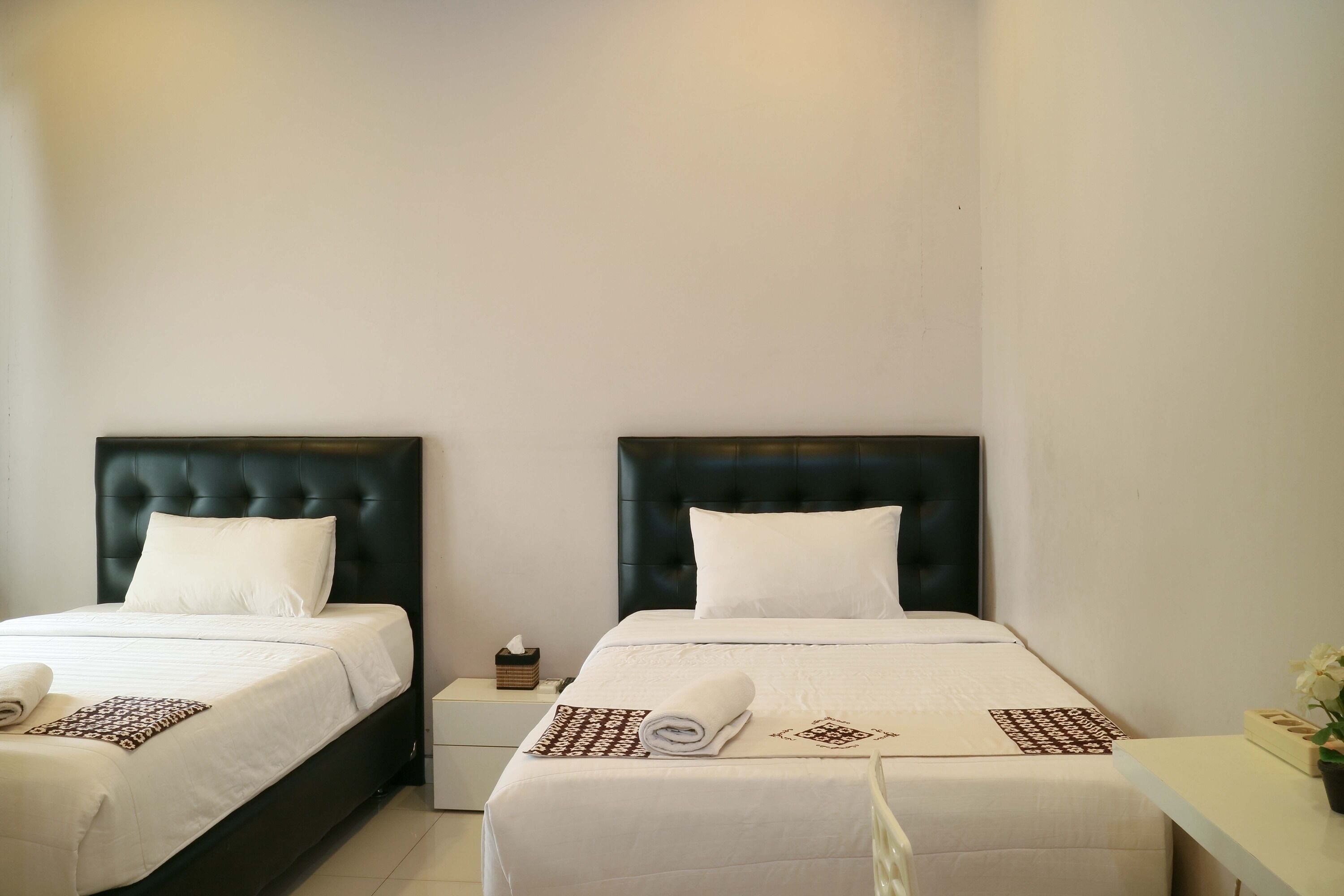 A hotel room with two beds, each with a black headboard and white bedding. There is a small bedside table with a tissue box and a rolled towel on each bed.