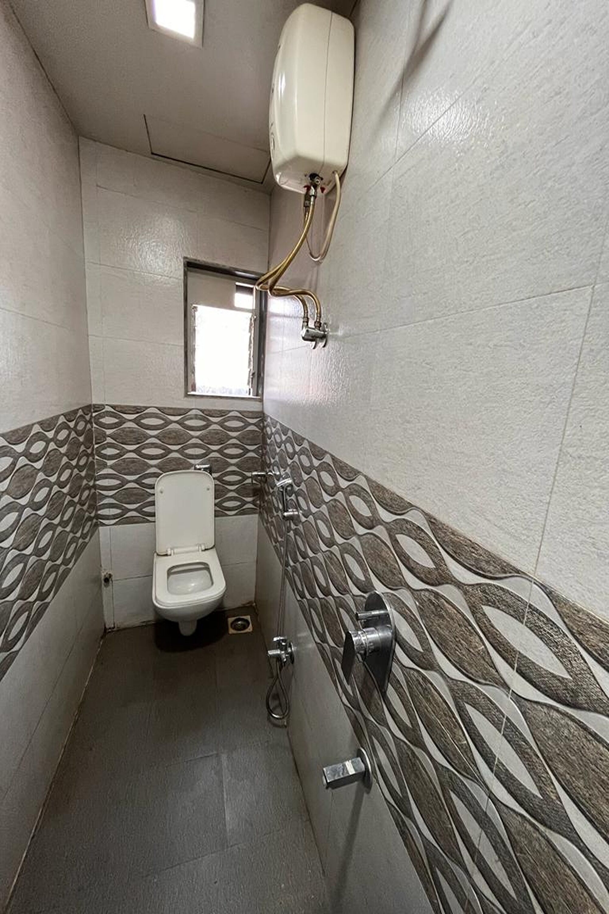 A small, narrow bathroom with patterned tiles, a toilet, a shower, and a window.