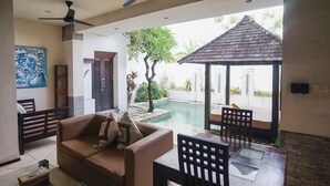 Uma sala de estar com sofá, mesa, cadeiras, ventilador de teto, um quadro e vista para uma piscina e área externa.
