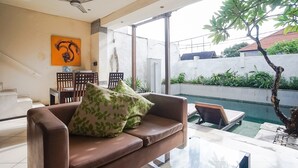 Sala de estar com sofá marrom, almofadas estampadas verdes, ventilador de teto e vista para uma piscina e vegetação lá fora.