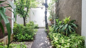 Um caminho estreito ladeado por vegetação exuberante e um muro de concreto, que conduz a um edifício branco.