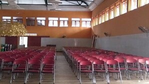 Une grande salle avec des rangées de chaises rouges, un plafond haut avec des poutres métalliques et des fenêtres le long des murs.