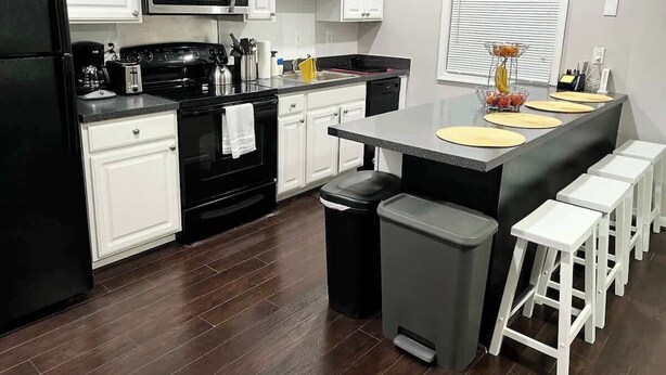 A modern kitchen with white cabinets, a black refrigerator, a microwave, and a stove. There are bar stools and a trash can.