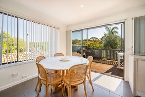 A dining area with a round table and four wicker chairs, a sliding glass door leading to an outdoor deck, and a view of greenery outside. - "Family Tides" – Short Stroll to the Beach (Copacabana)