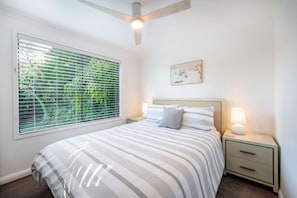 A bedroom with a bed, a nightstand, a wall-mounted lamp, a ceiling fan, and a window with blinds. - "Family Tides" – Short Stroll to the Beach (Copacabana)
