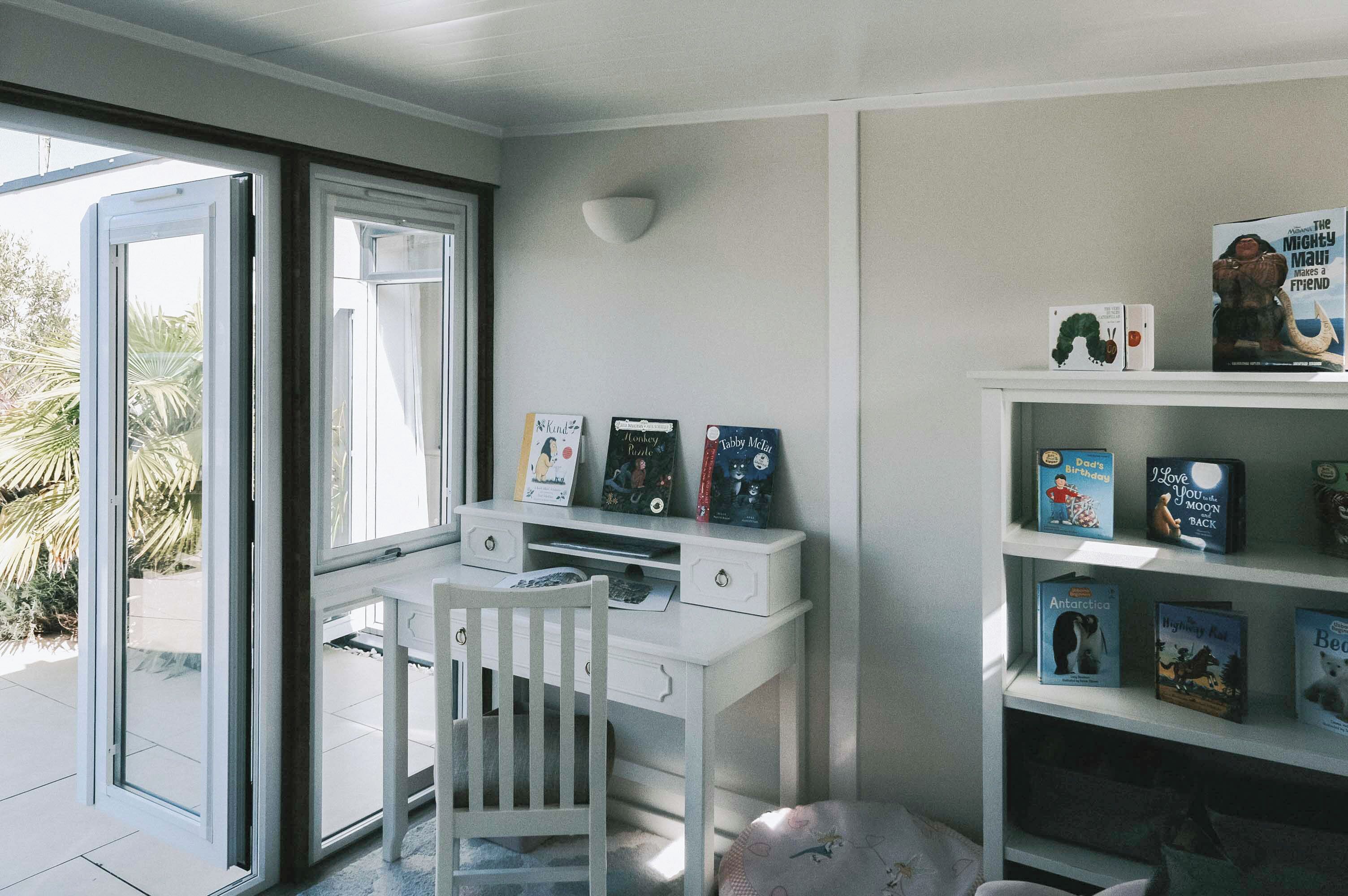 Ein Zimmer mit einem weißen Schreibtisch, einem Stuhl, einem Bücherregal mit Büchern und einem Fenster mit Blick auf Palmen.