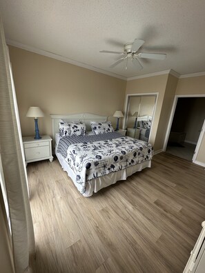 Un dormitorio con cama, dos mesitas de noche, un ventilador de techo y una puerta que da a otra habitación.