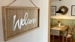 Un letrero de bienvenida de madera con diseño rústico, un área de comedor con mesa y sillas, y una guirnalda en la pared.