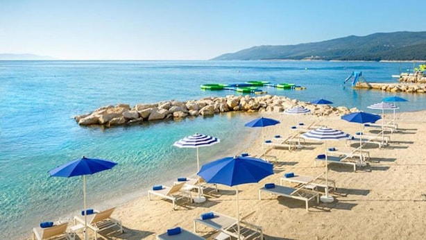 A beach with clear blue water, white lounge chairs, and blue umbrellas. - Stylish Villa with Pool (Labin)