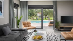 A modern living room with a grey sofa, a beige armchair, and a coffee table with lemons. A TV is mounted on the wall, and a pool is visible through the glass doors. - Stylish Villa with Pool (Labin)