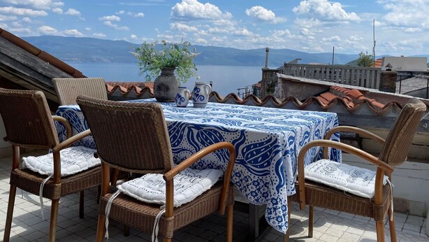 A terrace with wicker chairs and a table covered with a blue and white patterned tablecloth, overlooking a body of water and mountains. - Pleasant holiday home in Vrbnik near Secret Beach (Vrbnik)