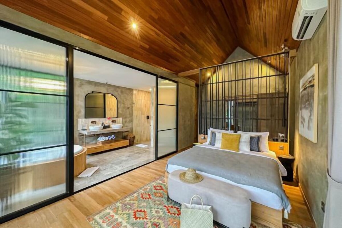 A bedroom with a large bed, a wooden ceiling, a sliding glass door leading to a bathroom, and a small bedside table.