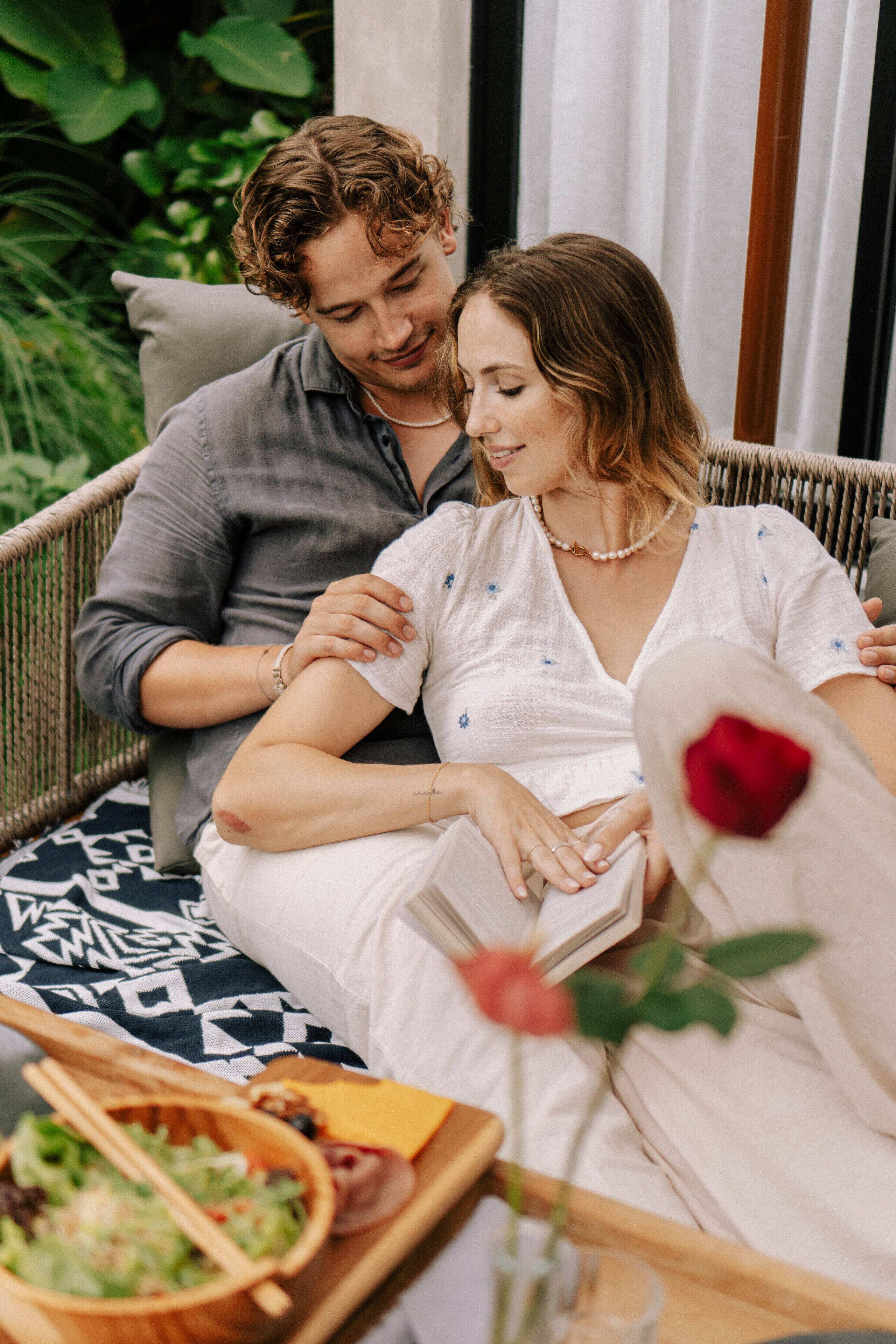 A couple sitting on a wicker sofa with a tray of food and a rose in a vase.
