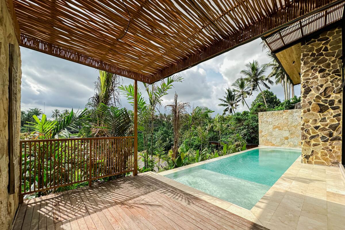 A wooden deck with a pool, surrounded by lush greenery and a stone wall.