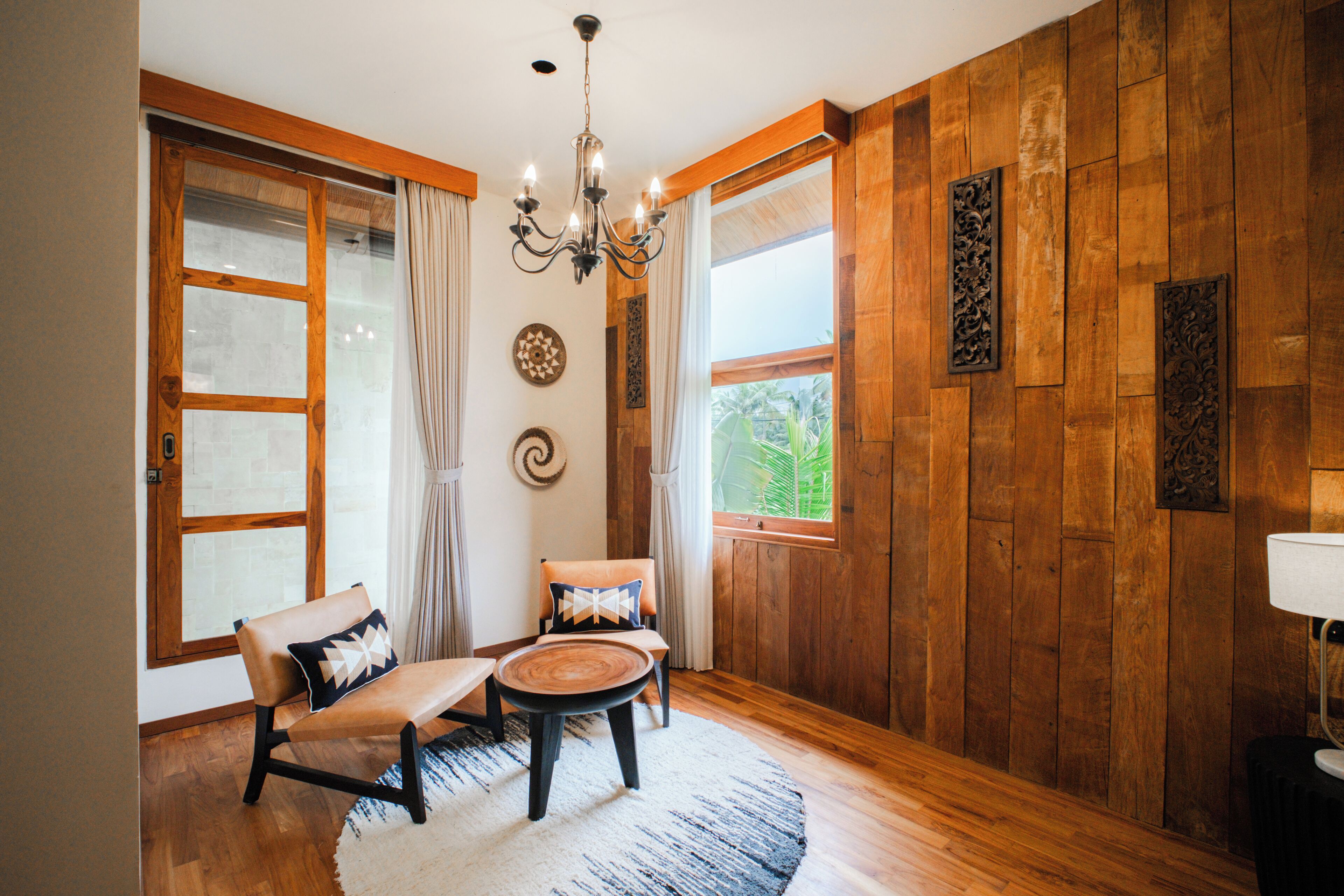 Une pièce aux murs en bois, ornée d’un lustre, comprenant deux canapés avec des coussins, une table basse ronde et une fenêtre avec des rideaux.