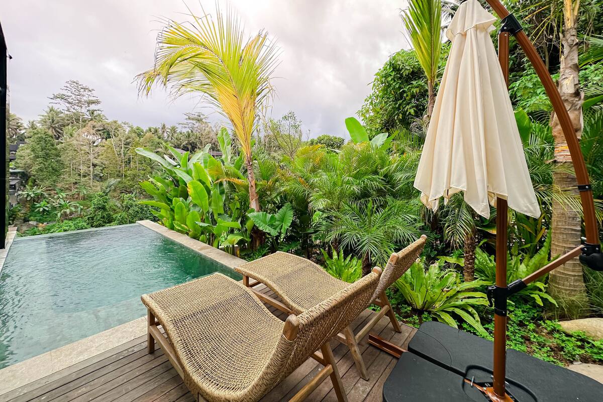 A poolside area with wicker lounge chairs, a wooden deck, and lush tropical vegetation.