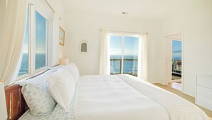 A bedroom with a large bed, a nightstand, a window with curtains, a small picture frame, and a view of the sea.