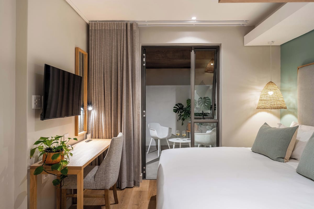 A hotel room with a bed, a desk, a chair, a TV, a plant, a sliding door, and a table with a glass top.