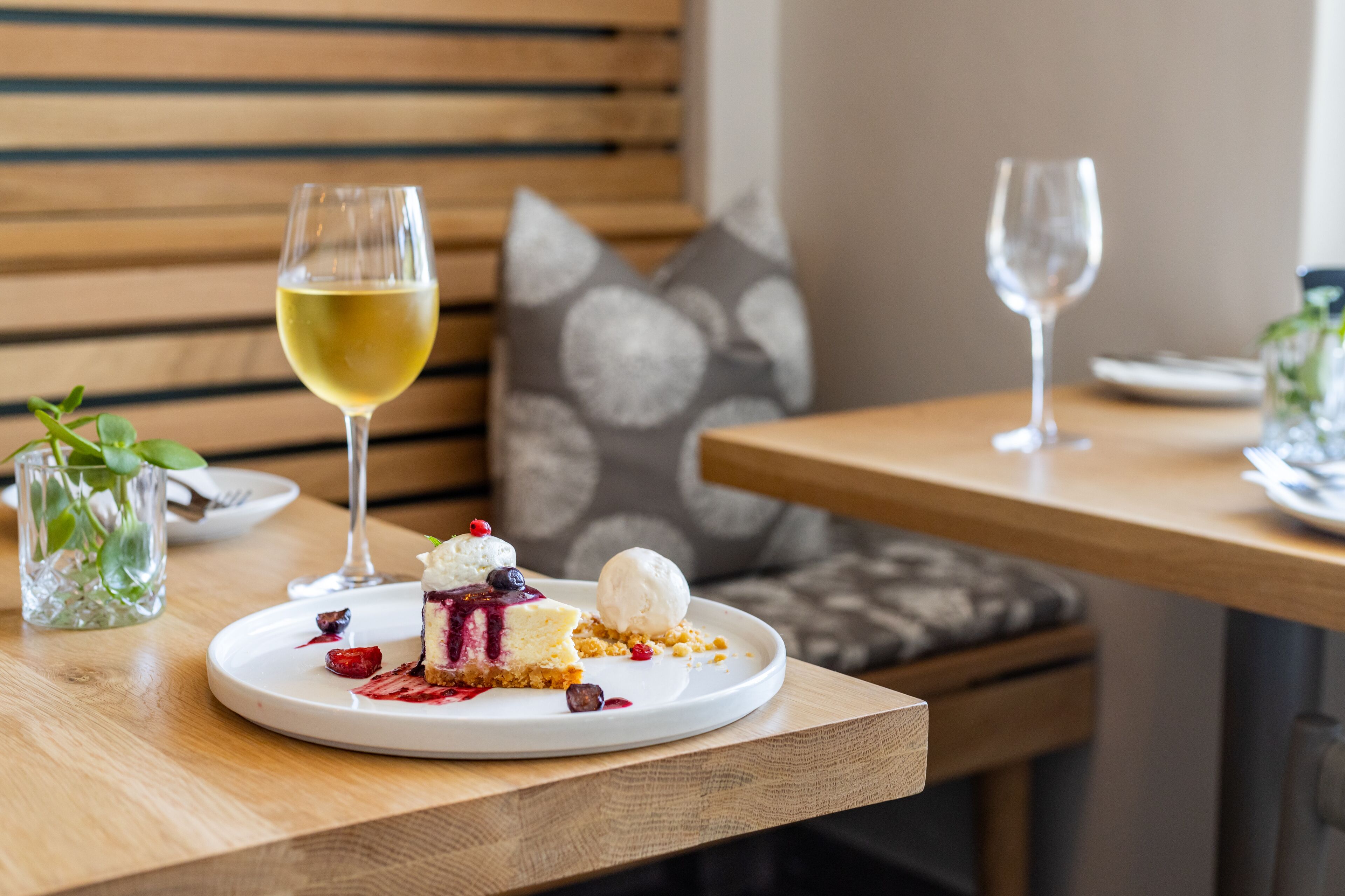A dining table with a dessert plate containing a slice of cake with berries and a scoop of ice cream, accompanied by a glass of white wine.