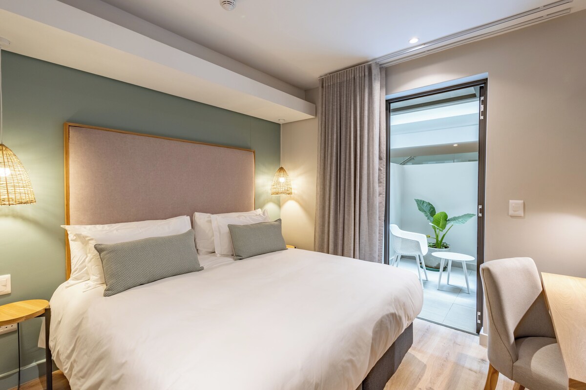 A modern hotel room with a large bed, bedside lamps, a small table, and a view of the outdoors.