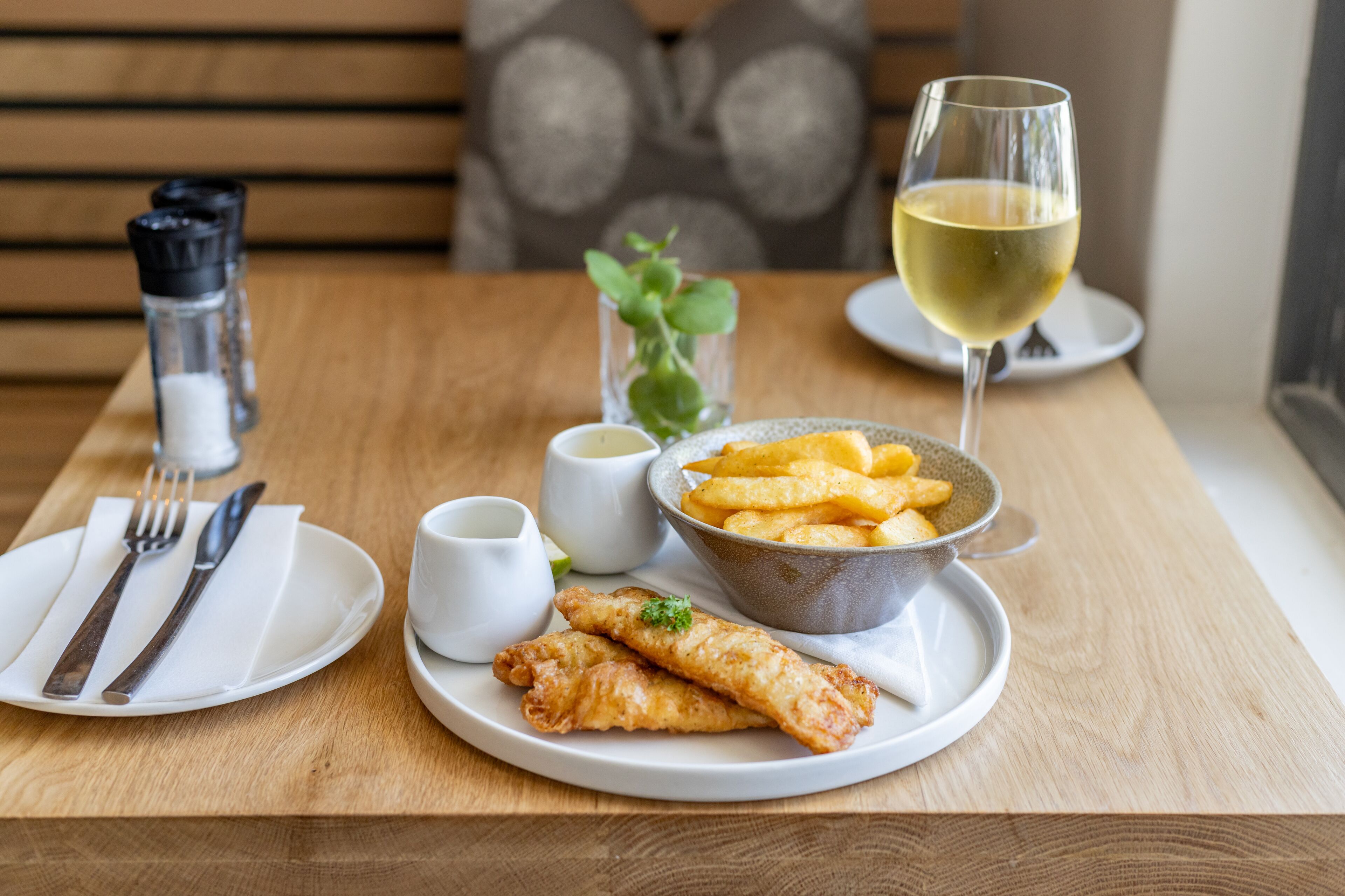 Una mesa de comedor con un plato de pescado y papas fritas, un bol de papas y un vaso de vino blanco, junto con salsas.