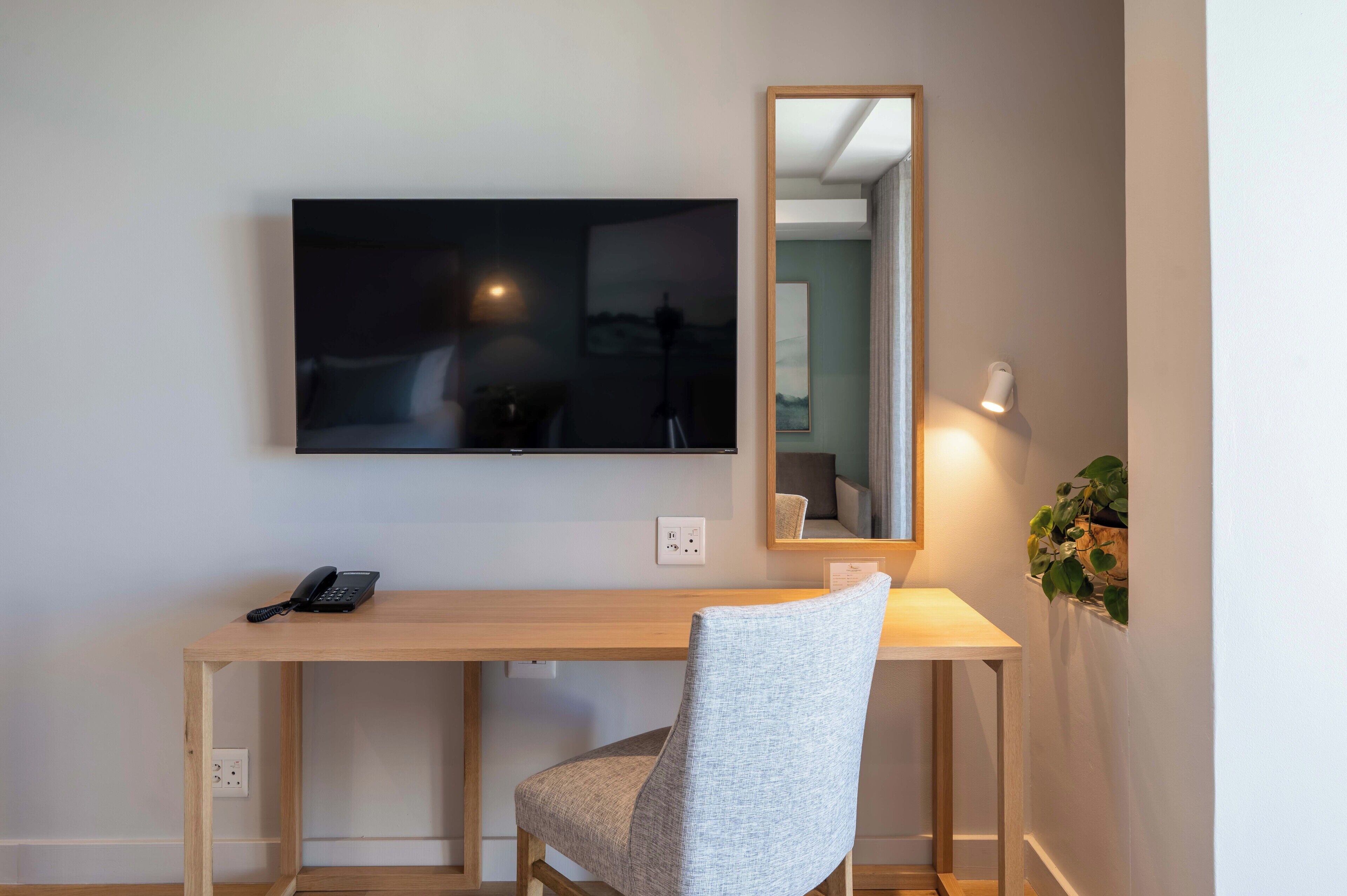 A room with a wooden desk, a chair, a flat-screen TV mounted on the wall, a mirror, and a wall-mounted lamp.