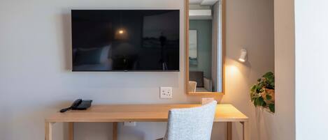 A room with a wooden desk, a chair, a flat-screen TV mounted on the wall, a mirror, and a wall-mounted lamp.