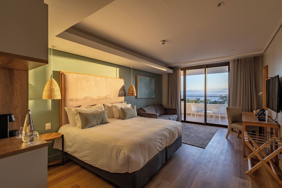 A modern hotel room with a large bed, a desk, a chair, and a balcony with a view.