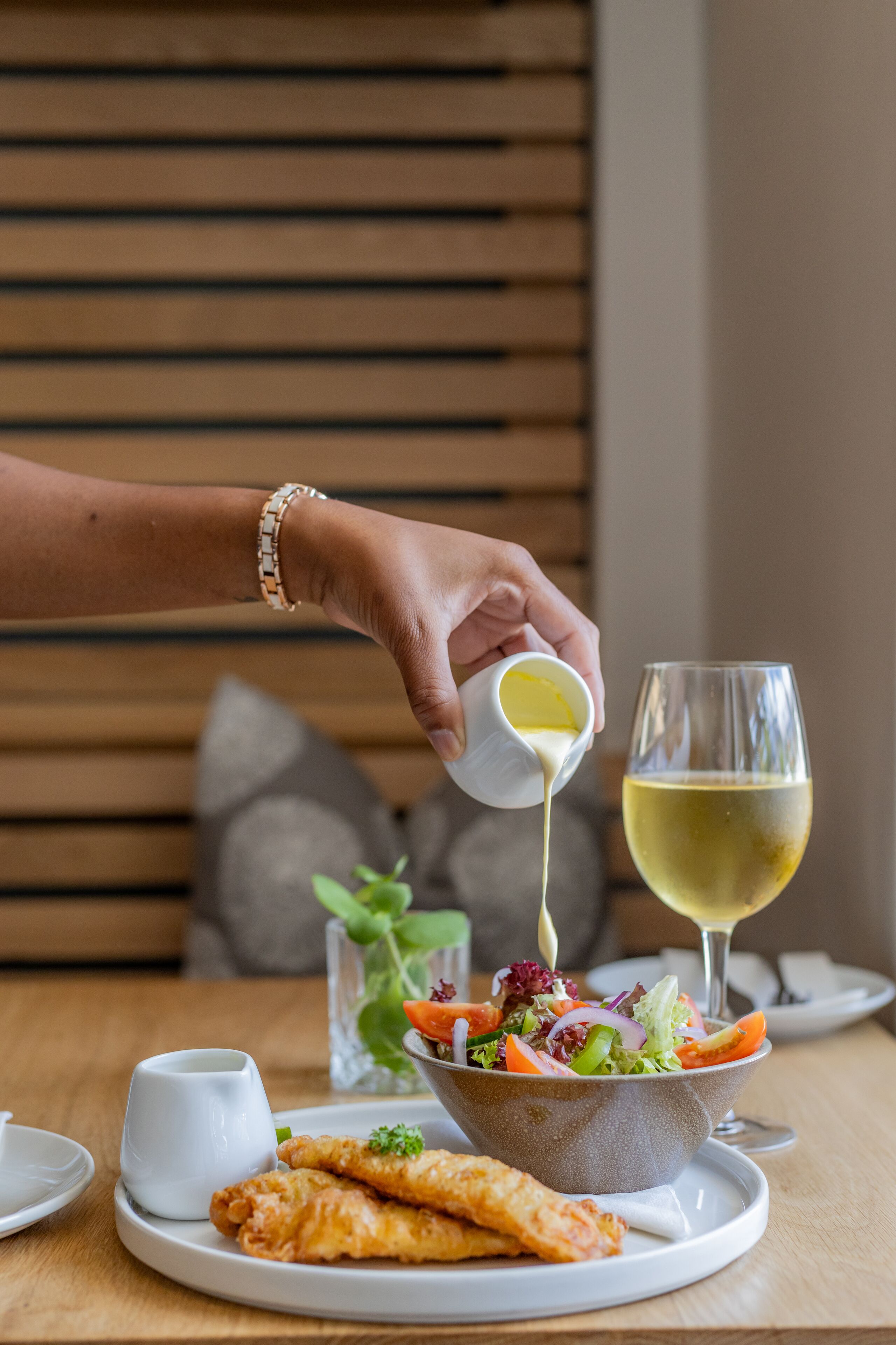 A hand pouring sauce into a glass of white wine, a bowl of salad, and fried items on a plate.
