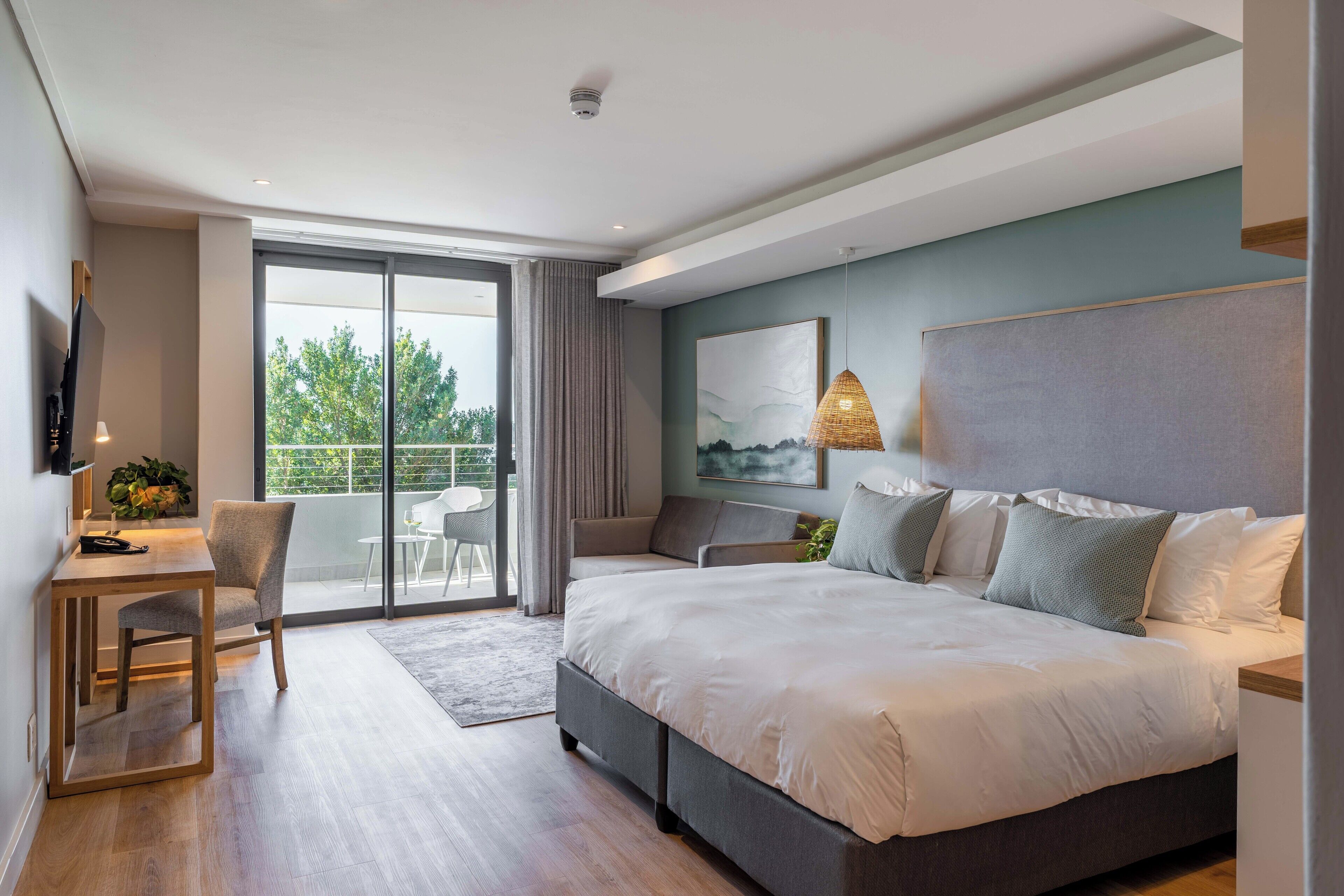 A modern bedroom with a large bed, a desk, and a chair. There is a balcony with a view of trees.