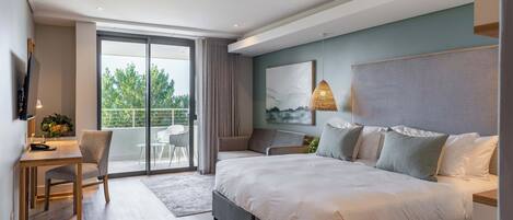A modern bedroom with a large bed, a desk, and a chair. There is a balcony with a view of trees.