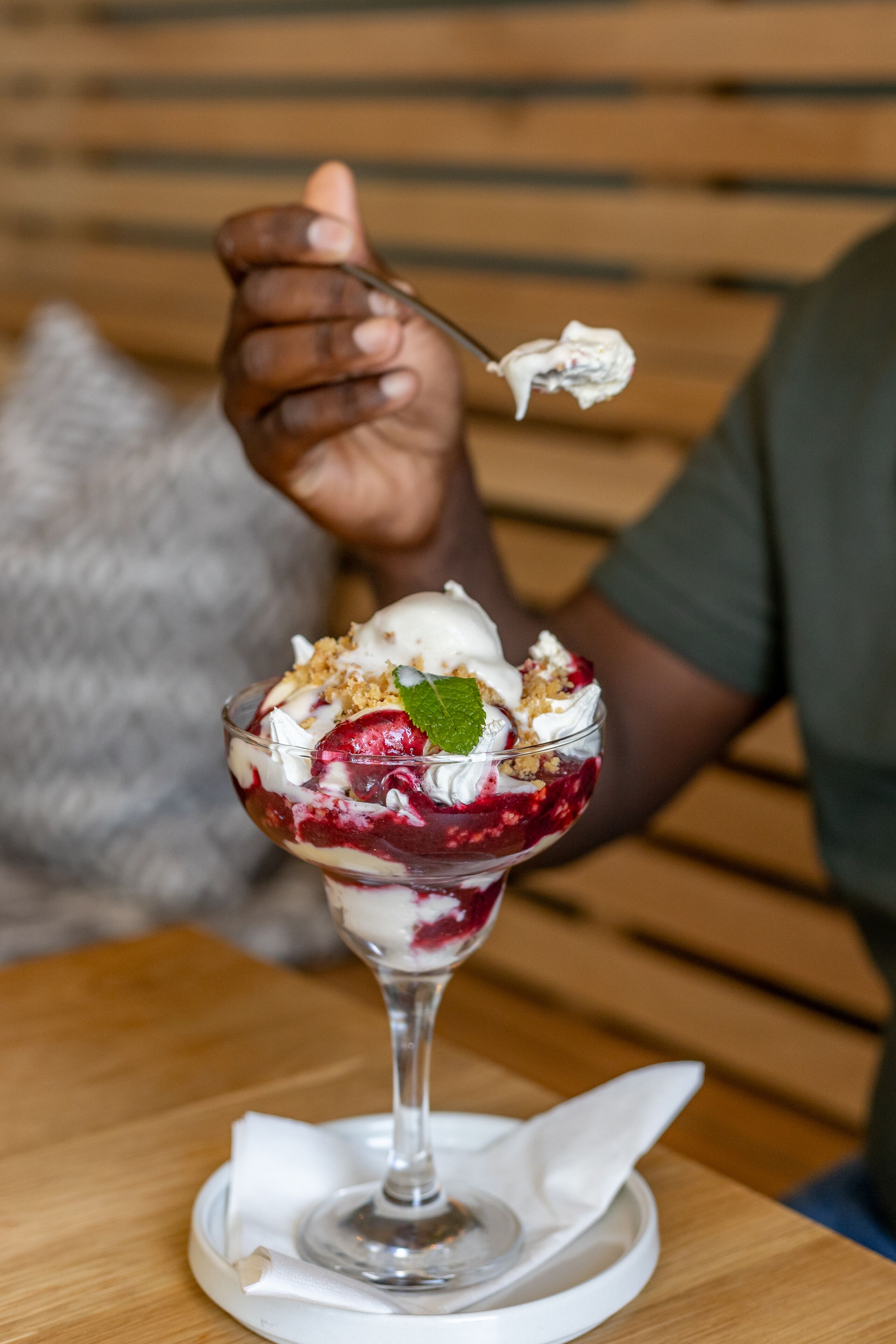 A dessert in a glass bowl with a spoon, placed on a white napkin on a wooden table.