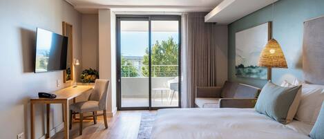 Habitación de hotel moderna con una cama grande, un escritorio con silla, televisión, balcón con vistas a la vegetación y un cuadro en la pared.