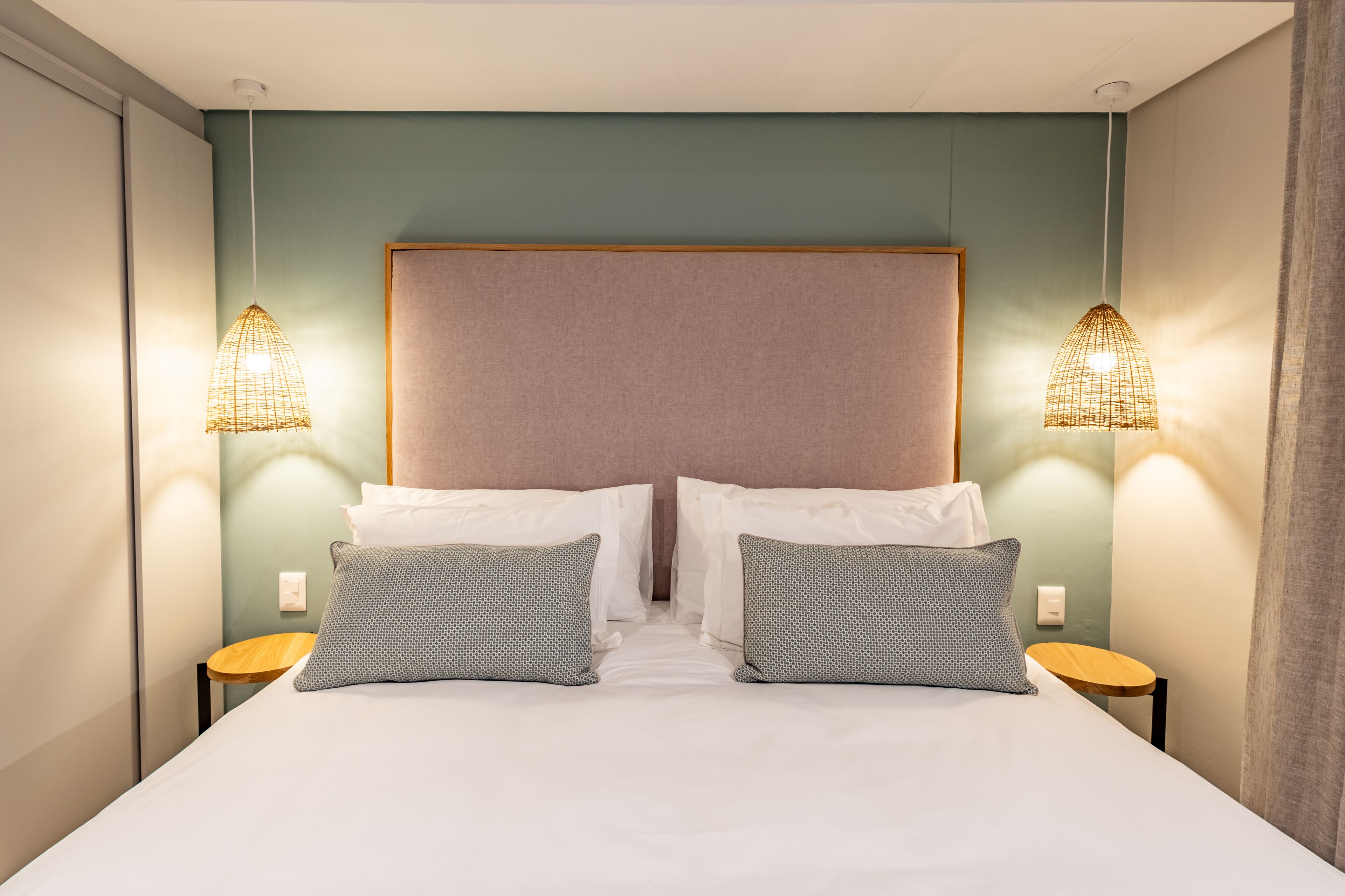 A double bed with white linens, two gray patterned pillows, and a wooden headboard. There are two bedside tables with wicker lamps on either side.