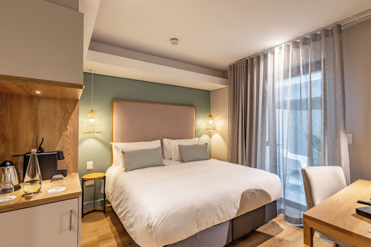Una habitación de hotel moderna con una cama grande, lámparas de noche, un escritorio de madera y vistas al exterior a través de cortinas traslúcidas.